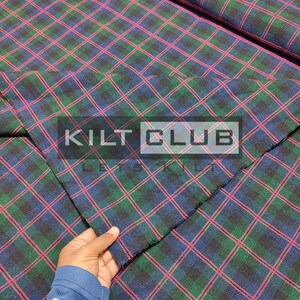 Könnte beinhalten: Nahaufnahme eines karierten Stoffes mit einem sich wiederholenden Muster aus grünen, marineblauen und roten Linien. Der Stoff wird von einer Hand gehalten. Der Text "KILT CLUB" ist auf dem Stoff überlagert.
