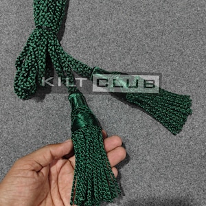 May include: A dark green kilt accessory with a braided cord and two tassels. The text "KILT CLUB" is visible on the image. The tassels are made of tightly wound threads, and the cord is knotted at the top.