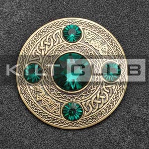 Puede incluir: Un broche redondo en oro antiguo, con intrincados diseños celtas y cinco gemas verde esmeralda. El broche presenta un diseño detallado con patrones intrincados, sobre un fondo gris oscuro.