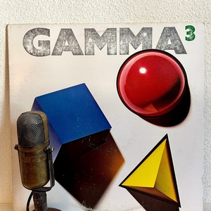 May include: Vintage microphone in front of a record album cover. The album cover features the word "GAMMA³" in distressed gray lettering, with a red sphere, blue cube, and yellow triangle.