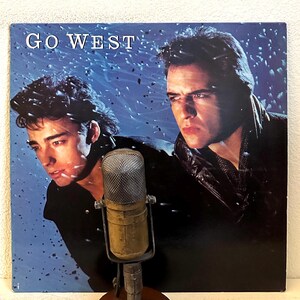 May include: A vintage vinyl record album cover featuring two men in black leather jackets against a blue and white background with a snow effect. The album title "GO WEST" is printed in white letters at the top of the cover.