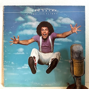 May include: A vintage vinyl record album cover featuring a man in a purple shirt and white pants jumping in the air against a blue sky with white clouds. The album title is "Endless Flight" by Leo Sayer.