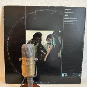 May include: A retro brass microphone is positioned in front of a vinyl record album. The album cover is black with a photograph of people and a list of song titles including "Good Morning" and "I Love You". The microphone has a mesh screen and a round base.