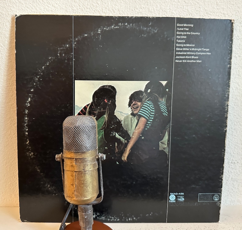 May include: A retro brass microphone is positioned in front of a vinyl record album. The album cover is black with a photograph of people and a list of song titles including "Good Morning" and "I Love You". The microphone has a mesh screen and a round base.