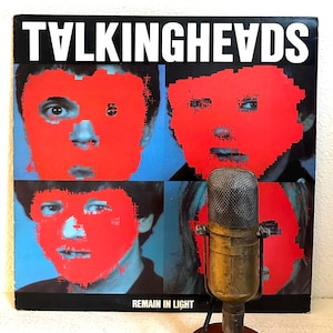 May include: Album cover for Talking Heads' "Remain in Light" featuring four red-pixelated faces on a blue background, with the band's name in white at the top. An antique microphone is in the foreground.