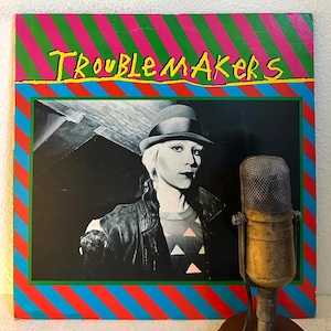 May include: Vintage vinyl record album cover titled "TroubleMakers". The cover features a black and white photo of a person wearing a hat and jacket. A vintage microphone is in the foreground. The album art has a colorful striped border.