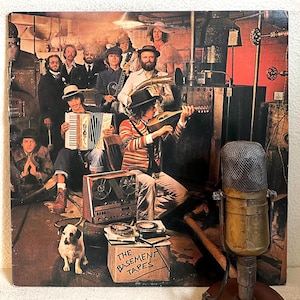 May include: Album cover for "The Basement Tapes" featuring a group of musicians with various instruments. The cover art shows the band in a dimly lit room with a dog, a vintage microphone, and a reel-to-reel tape recorder.