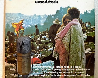 Puede incluir: Micrófono vintage frente a una portada del álbum Woodstock. La portada muestra una multitud, una cometa colorida y el texto "Woodstock". El álbum es un conjunto de 3 discos.