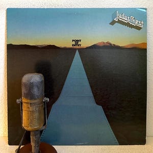 May include: Judas Priest "Point of Entry" album cover. The album art features a blue road leading to the horizon under a blue and orange sky. The band's name is in a silver, angular font. A vintage microphone is in the foreground.