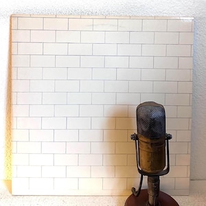 May include: A square vinyl record cover featuring a white brick wall pattern with thin gray mortar lines. Positioned in front of the lower right corner is a vintage-style microphone with a metallic mesh head and a cylindrical body on a stand.