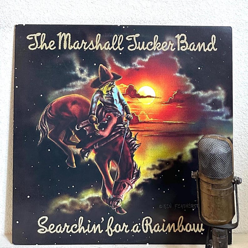 May include: A black vinyl record album cover with a cowboy riding a bucking horse in front of a sunset. The album cover has the text 'The Marshall Tucker Band' in gold lettering at the top and 'Searchin' for a Rainbow' in gold lettering at the bottom. The album cover is in front of a microphone.