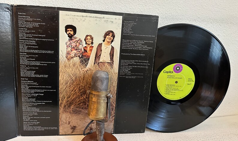 May include: An album cover featuring The Steve Miller Band with a vintage microphone in the foreground. The album cover shows the band members standing in tall grass. The vinyl record is black with a yellow and purple label.