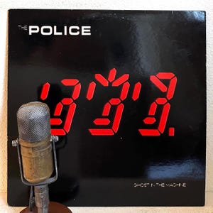 May include: A vintage microphone stands before a black album cover. The album cover features the band name "THE POLICE" in white, with large red digital-style numbers. The album title "GHOST IN THE MACHINE" is at the bottom.