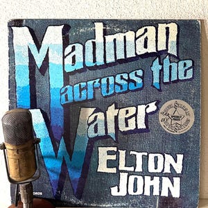 May include: A vinyl record album cover with a blue and white design. The album is titled "Madman Across the Water" by Elton John. The cover features a blue and white striped background with the album title in white letters. There is a sticker on the bottom right corner that says "Super Stars at Super Savings - Specially Priced".
