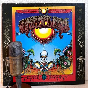 May include: A vintage microphone stands before a vinyl record album cover. The album art features a vibrant design with a skull, sun, and intricate lettering against a blue sky and red border. The album is titled "Grateful Dead."