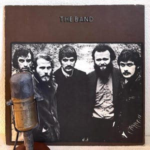 May include: Vintage album cover for "The Band" with a brown background and a black and white photo of the band members. A vintage microphone is in the foreground. The album title is in silver letters.