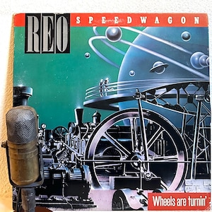 May include: A vintage vinyl record album cover featuring the band REO Speedwagon. The cover art is a futuristic illustration of a large, silver spaceship with a green background. The album title "Wheels are turnin'" is printed in white at the bottom of the cover.