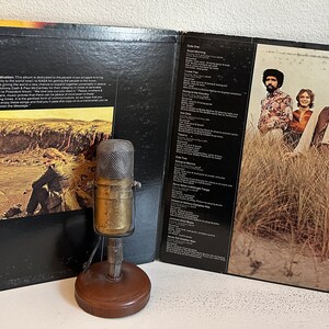 May include: Vintage vinyl record album with a black cover and a sepia-toned photo of a person on a hillside. The album is open, revealing song lyrics and a photo of a band. An antique microphone sits in front.