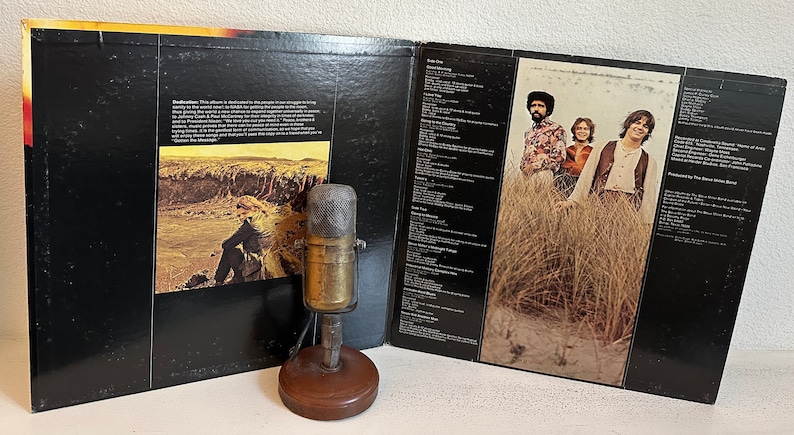 May include: Vintage vinyl record album with a black cover and a sepia-toned photo of a person on a hillside. The album is open, revealing song lyrics and a photo of a band. An antique microphone sits in front.