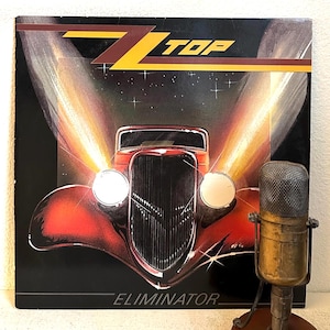 May include: Album cover for ZZ Top's Eliminator, featuring a red hot rod with bright headlights and the band's logo in yellow. A vintage microphone is in the foreground, suggesting a music theme.