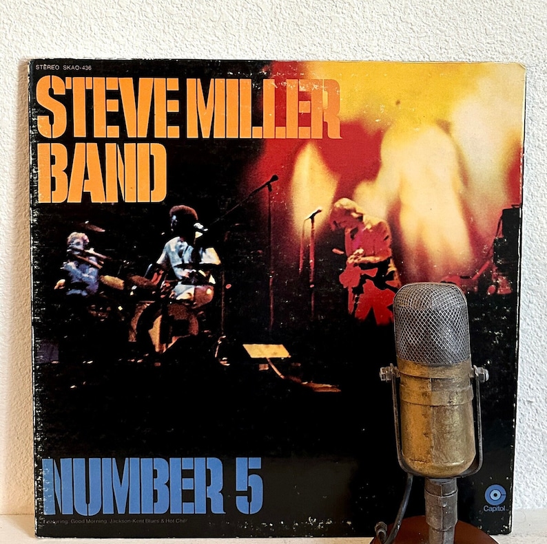 May include: A vintage vinyl record album cover for the Steve Miller Band's "Number 5". The album cover features the band performing on stage with the band's name in orange block letters. A vintage microphone is in the foreground.