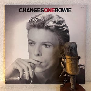 May include: Album cover for David Bowie's "ChangesOneBowie" with a black and white portrait of the artist. A vintage microphone is in the foreground. The album title is in black and red text.