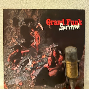 May include: A vintage vinyl record album cover featuring a band called Grand Funk Railroad. The album cover is a dark, cave-like setting with four people dressed in primitive clothing. The album title "Survival" is written in red letters.