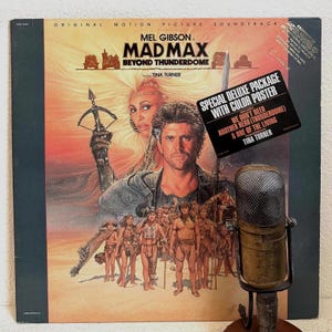 May include: A vintage vinyl record album cover for the movie "Mad Max Beyond Thunderdome" featuring Mel Gibson and Tina Turner. The cover art is a movie poster with a desert scene and the film's title. A sticker reads "Special Deluxe Package with Color Poster."