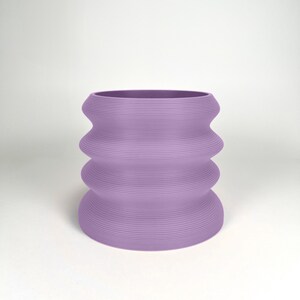 May include: A lavender-colored ceramic planter with a wavy, ribbed design. The planter has a wide, round opening and a slightly narrower base.