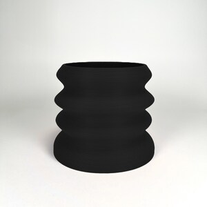 Könnte beinhalten: Eine schwarze Keramikvase mit einem wellenförmigen, geriffelten Design. Die Vase ist hoch und zylindrisch.