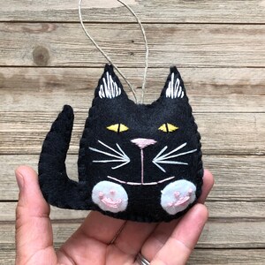 Op de afbeelding: Zwarte vilt katten ornament met witte geborduurde snorharen en gele ogen. De kat heeft roze vilt poten.
