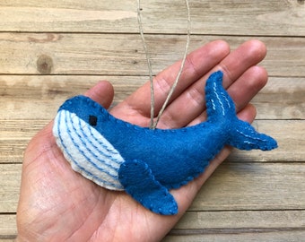 May include: A handmade blue felt whale ornament with white accents and a string for hanging. The whale features stitched details and is held in a hand against a wooden background. A charming nautical-themed decoration.