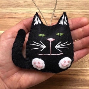 Op de afbeelding: Zwarte vilt katten ornament met witte geborduurde snorharen en groene ogen. De kat heeft roze vilt poten.