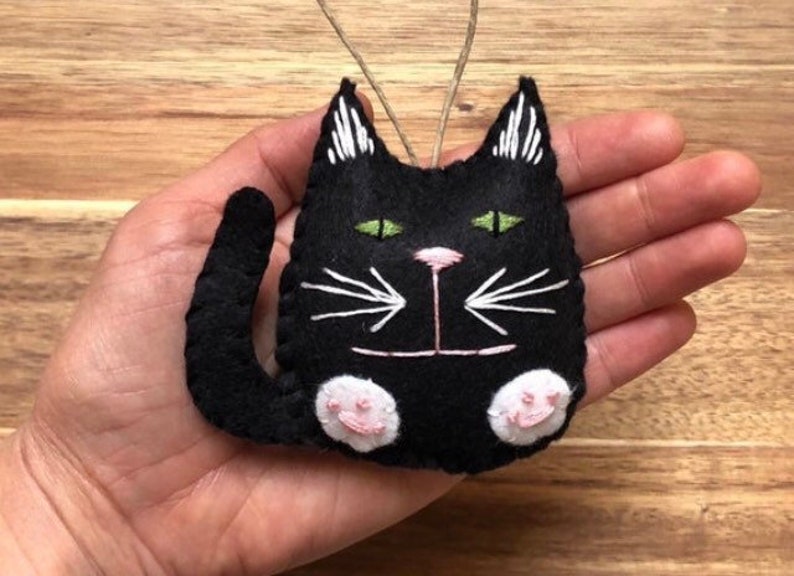 Op de afbeelding: Zwarte vilt katten ornament met witte geborduurde snorharen en groene ogen. De kat heeft roze vilt poten.