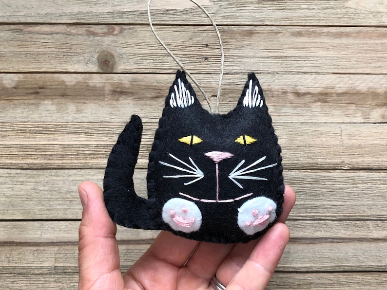 Op de afbeelding: Zwarte vilt katten ornament met witte geborduurde snorharen en gele ogen. De kat heeft roze vilt poten.