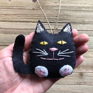 Op de afbeelding: Zwarte vilt katten ornament met geborduurde witte snorharen, gele ogen en roze neus. De kat heeft een lange staart en witte poten.