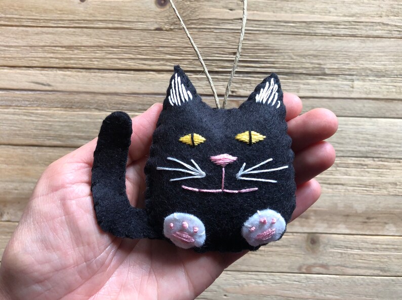 Op de afbeelding: Zwarte vilt katten ornament met geborduurde witte snorharen, gele ogen en roze neus. De kat heeft een lange staart en witte poten.