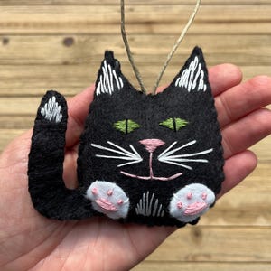 Op de afbeelding: Zwarte vilt kattenversiering met witte geborduurde details, waaronder pootjes, snorharen en een neus. De kat heeft groene ogen en een roze neus.