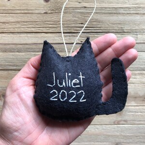 Op de afbeelding: Zwarte viltkattenversiering met witte borduurwerk dat "Juliet 2022" zegt.