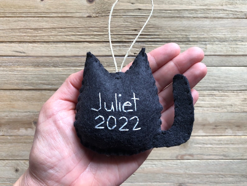 Op de afbeelding: Zwarte viltkattenversiering met witte borduurwerk dat "Juliet 2022" zegt.