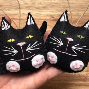 Op de afbeelding: Twee zwarte viltkatten ornamenten met geborduurde details. E&eacute;n kat heeft gele ogen en de andere heeft groene ogen. Beide katten hebben een roze neus en witte snorharen.