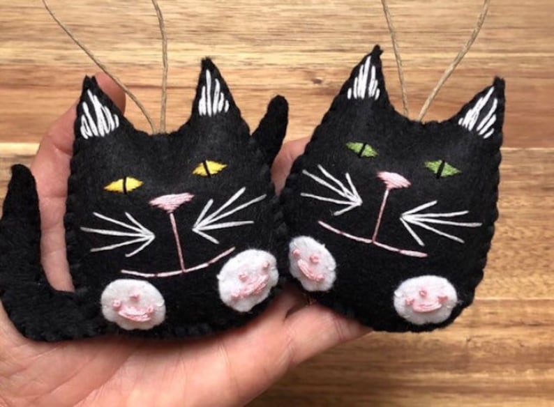 Op de afbeelding: Twee zwarte viltkatten ornamenten met geborduurde details. E&eacute;n kat heeft gele ogen en de andere heeft groene ogen. Beide katten hebben een roze neus en witte snorharen.
