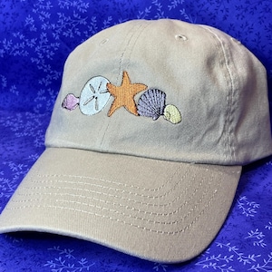 May include: Tan baseball cap with embroidered seashells, including a sand dollar, starfish, and various shells in shades of orange, purple, and yellow. The cap has a curved brim and a stitched design.