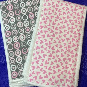 May include: Two baby burp cloths. One cloth has a pattern of pink, gray, and white circles on a gray background. The other cloth has a pattern of pink hearts on a white background. Both cloths have white terry cloth borders.