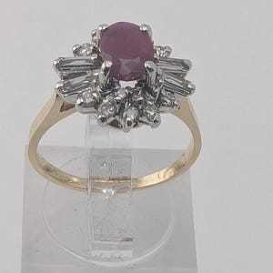 Peut inclure: Une bague en or avec une pierre précieuse ovale rose foncé au centre, entourée d'un groupe de petits diamants ronds et de diamants taille baguette sertis dans une monture en métal blanc, avec un anneau en or jaune poli.