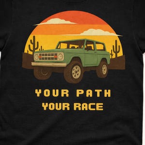 Puede incluir: Camiseta negra con un gráfico de un SUV verde vintage sobre un fondo de puesta de sol desértica con cactus. El texto "YOUR PATH YOUR RACE" está impreso en amarillo debajo del vehículo.