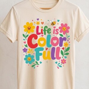 以下が含まれることがあります： 「Life is Color Full」という太字のカラフルな文字がプリントされたクリーム色のTシャツ。デザインには、赤、黄、ピンク、青の鮮やかな色合いで、笑顔の花、小さなハート、飛んでいるハチが描かれています。