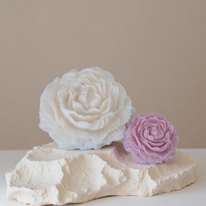 Könnte beinhalten: Zwei blütenförmige Kerzen, eine große weiße und eine kleinere lavendelfarbene, liegen auf einem strukturierten, cremefarbenen Stein. Die Kerzen sind mit Blütenblattformationen detailliert, was ein Blumendesign ergibt.