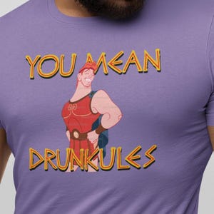 May include: A lilac-colored t-shirt featuring a cartoon illustration of Hercules with the text "YOU MEAN DRUNKULES" in yellow and orange. The graphic shows Hercules in a red tunic and blue cape, smiling.