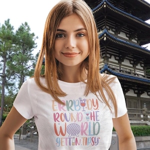 May include: White t-shirt with the text "Everybody Round the World Gettin Tipsy" in colourful letters. The shirt features a cocktail and a globe graphic. The model is smiling in front of a Japanese-style building.